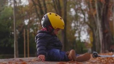 Genç bir çocuk, sakin bir yolda, düşmüş yaprakların arasında oturur. Çevrenin küçük elementlerini alıp, açık havada sakin bir keşif anı yaratırken dikkatle bakar.