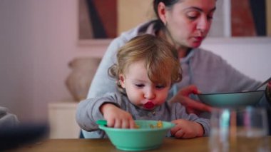 Çocuk kaşıkla kendini beslemeye konsantre olurken, bakıcı kendi yemeğinin yanında kalır ve paylaşılan evde özerklik ve özerklik geliştirdiğini gösterir.