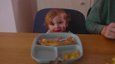 Genç çocuk tabağın üzerinden bakıp gülümsüyor, tanıdık bir ev ortamında rahatça yemek yerken merak ve rahatlık gösteriyor.