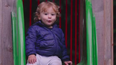 Yeni yürümeye başlayan çocuk, parkta oyun parkından kayarak kayıyor, sıcak bir palto giyerek, heyecan ve merakını ifade ederken, bir yandan da dışarıda eğleniyor.