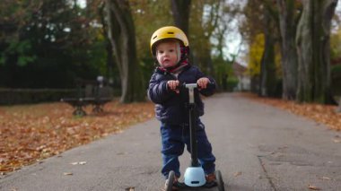 Küçük bir çocuk elinde bir scooter tutarken duraksar, sessiz bir merakla görünmeyen bir şeye bakar sıcak sonbahar renkleri ve dağınık yapraklar yumuşak mevsimsel bir atmosfer yaratır.