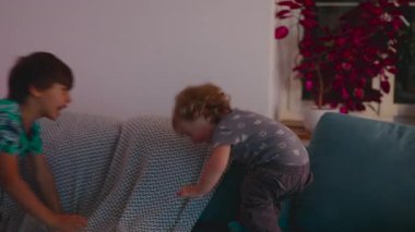 Büyük çocuk kanepede yatarken, küçük çocuk yan taraftaki minderlere uzanır. Örtülü bir battaniye, mobilya ve oturma odasında görülebilir ev eşyalarıyla.