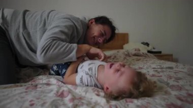 Yetişkin ve küçük çocuklar yatakta neşeli bir etkileşim içinde, kahkaha, şefkat, güvenlik, duygusal yakınlık, güven ve neşeli aile ilişkilerini ifade ediyorlar.