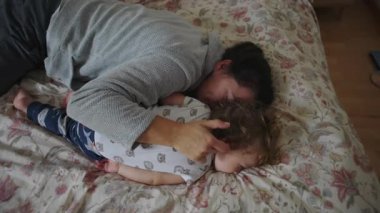 Anne, bebeğini yatakta nazikçe kucaklıyor. Bakım, güvenlik, duygusal güvenlik, bağlılık ve evdeki samimi aile hayatını ifade ediyor.