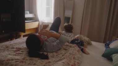 Bebek yatağın etrafında heyecanlı bir şekilde hareket ederken anne gülümseyerek ve nazikçe oynaşarak rahat bir yatak odası ortamında otantik bir bağlantı anı gösteriyor.