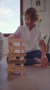 Çocuk dikkatli bir şekilde yüksek kuleye blok ekler, kardeş gözlemleri ise risk alarak odak koordinasyonu sağlar ve neşeli ev ortamında heyecanı paylaşır