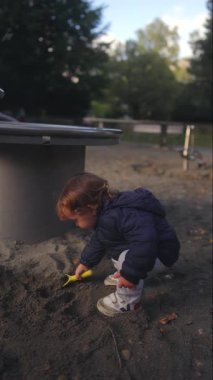 Küçük çocuk oyuncak kürek kullanarak kumu transfer ediyor. Odaklanmış keşifleri, motor gelişimini ve oyun alanındaki keşifleri ifade ediyor.