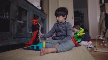 Büyük çocuk oyuncak setini birbirine bağlarken, yeni yürümeye başlayan çocuk yakınlarda kardeşlerinin birlikte yaşama merakını ve evde günlük oyun rutinini ifade ediyor.