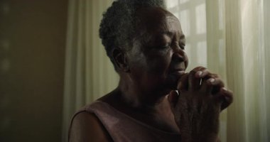 Kıdemli kadın evde dua ederken elini sıkar, bağlılık, huzur, kabullenme, umut ve ebedi iç huzuru imanla ifade eder.