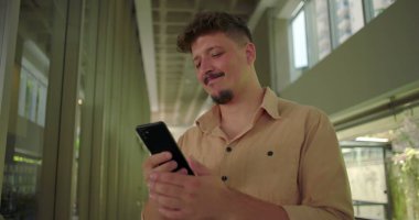 Ofis koridorunda akıllı telefonu tutan genç adam, ekrandaki mesaja olumsuz tepki veriyor, profesyonel iş ortamında hayal kırıklığına uğramış ifade