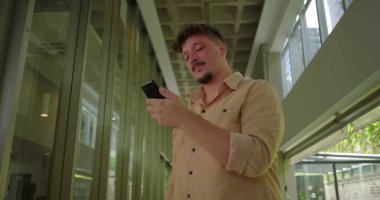 Ofis koridorundaki genç adam akıllı telefona bakarken hayal kırıklığıyla tepki veriyor. Profesyonel ortamdaki hayal kırıklığına uğramış yüz ifadesi.