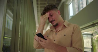 Ofis koridorunda akıllı telefonu tutan genç adam, ekrandaki mesaja olumsuz tepki veriyor, profesyonel iş ortamında hayal kırıklığına uğramış ifade
