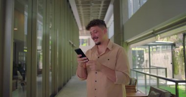 Genç adam telefonu tutuyor ve ekranda gülümsüyor. Profesyonel ofis koridorunda neşeli anlar yaşıyor. Sıradan ve dostane bir atmosfer.