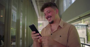 Genç adam telefonu tutuyor ve ekranda gülümsüyor. Profesyonel ofis koridorunda neşeli anlar yaşıyor. Sıradan ve dostane bir atmosfer.