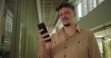 Ofis koridorunda akıllı telefonu tutan genç adam, ekrandaki mesaja olumsuz tepki veriyor, profesyonel iş ortamında hayal kırıklığına uğramış ifade