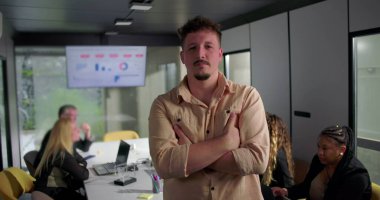 Kollarını kavuşturmuş genç adam ofis ortamında kararlı görünüyor, iş arkadaşları ekranda veri görselleştirmesi sırasında arka planda çalışıyor