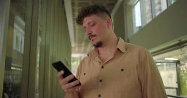 Ofis koridorunda akıllı telefonu tutan genç adam, ekrandaki mesaja olumsuz tepki veriyor, profesyonel iş ortamında hayal kırıklığına uğramış ifade