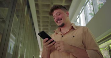 Ofis koridorunda akıllı telefona gülümseyen genç adam, mobil cihaz kullanırken neşeyle tepki veriyor, modern iş ortamında rahat ve neşeli bir an.