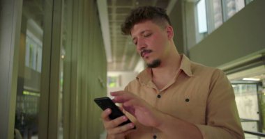 Ofis koridorunda akıllı telefonu tutan genç adam, ekrandaki mesaja olumsuz tepki veriyor, profesyonel iş ortamında hayal kırıklığına uğramış ifade