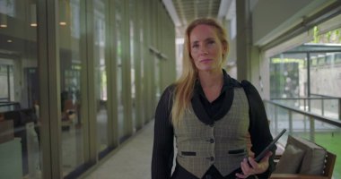 Modern koridorda elinde tabletle dikilen kadın, kameraya objektif bir ifadeyle bakıyor, profesyonel görev ya da buluşma için hazırlanıyor.