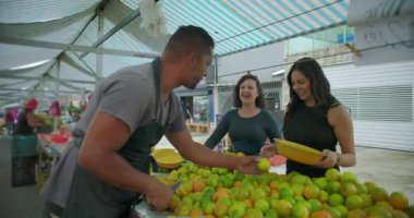 Hispanik satıcı, açık hava pazarında alışveriş yapanlarla sıcak temas kuruyor. Kişisel hizmet rutin etkileşimini ve toplum temelli gıda alışverişini ifade ediyor.