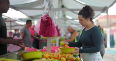 Kadın tüketici, mahalle pazarındaki sokak satıcısıyla ürün seçimini tartışıyor. Pratik etkileşim aşinalığını ve toplum temelli gıda alışverişini dile getiriyor.
