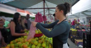 Mahalle pazarında taze ürünleri poşetleyen kadın satıcı günlük iş temposu ve insan ölçekli kamu gıda ticareti alışverişini dile getiriyor