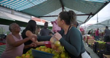 Kadın satıcı, mahalle pazarından alım yaptıktan sonra üst düzey müşteriyi selamlıyor. Dostluk, minnettarlık ve günlük insan ilişkilerini gıda ticaretiyle iletiyor.