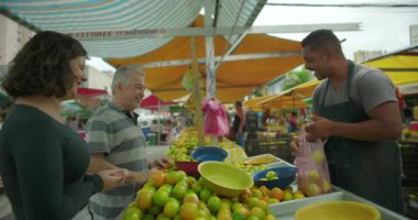 Genç kadın ve yaşlı erkek pazarda meyve seçerken, kısa bir sohbet, rutin market alışverişi ve yerel gıda ticaretine günlük katılımda bulunurken tedarikçiyle konuşuyor.