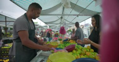 İspanyol satıcı taze meyveyle plastik poşet hazırlarken, kadınlar işbirlikçi alışveriş rutin etkileşimi ve mahalle piyasa hayatını yansıtan yakınlardaki narenciyi seçiyorlar.