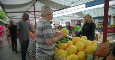 Dışarıdan taze meyve seçen kıdemli tüketici, rutin etkileşimi, karşılıklı saygıyı ve toplum bazlı sokak piyasa hayatı içinde yaşayan bağlantıyı iletiyor.