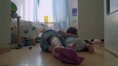 İki küçük kardeş, tahta zeminde güneşli yatak odasında birlikte oynuyorlar. Bebek, oyuncaklarla ve gün ışığıyla çevrili büyük çocuğu sıcak bir şekilde kucaklıyor.