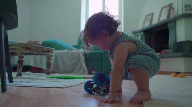 İçeride ahşap zemine çömelmiş küçük çocuk, yoğun merak ve denge ile mavi oyuncak robotu dikkatlice inceliyor, rahat oturma odası mobilyaları ile çevrili