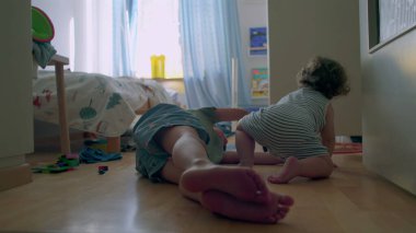 İki küçük kardeş, tahta zeminde güneşli yatak odasında birlikte oynuyorlar. Bebek, oyuncaklarla ve gün ışığıyla çevrili büyük çocuğu sıcak bir şekilde kucaklıyor.