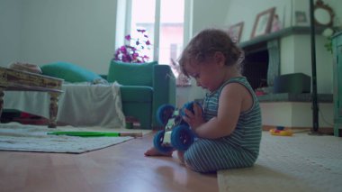 İçeride ahşap zemine çömelmiş küçük çocuk, yoğun merak ve denge ile mavi oyuncak robotu dikkatlice inceliyor, rahat oturma odası mobilyaları ile çevrili