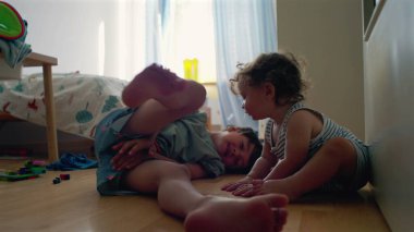 Yürüyen çocuk, büyük kardeşinin yatıp, yatak odasında oynamasını seyrediyor. Camdan gün ışığı akıyor. Etrafa saçılan renkli oyuncaklar.