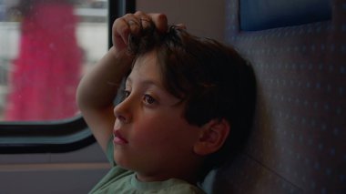 Çocuk sessizce tren penceresinden dışarı bakar, el şakağına, vücudunu koltuğa sıkıştırır, ciddi ve odaklanmış bir şekilde, manzara dışarı doğru koşarken.