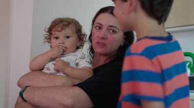 Anne kucağında bebek ve yanaktan nazikçe öpüşürken büyük çocuk yanında duruyor. Evin içinde özgün anne şefkati ve aile şefkati sahnesi.