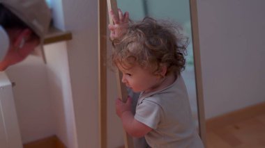 Küçük çocuk aynaya sarılıp kameraya doğru yürümeden önce arkasını dönüyor, kapalı alanda hareket, denge ve erken keşifler gösteriyor.