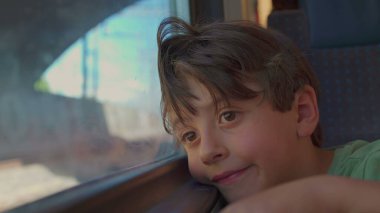 Genç çocuk trenin penceresinde dinleniyor, hüzünlü bir ifadeyle dışarı bakıyor, güneş ışığı yüzünü ve saçlarını aydınlatıyor gezinti sırasında manzara bulanıklaşırken