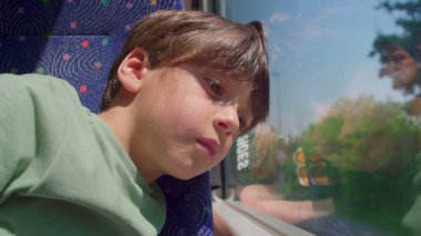 Genç çocuk yüzünü otobüs camına dayadı, gözler manzaranın önünden geçmeye odaklandı, güçlü yansıma dışarıdaki yoğun merakı ve doğal çevreyi ortaya çıkardı.