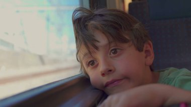 Genç çocuk trenin penceresinde dinleniyor, hüzünlü bir ifadeyle dışarı bakıyor, güneş ışığı yüzünü ve saçlarını aydınlatıyor gezinti sırasında manzara bulanıklaşırken