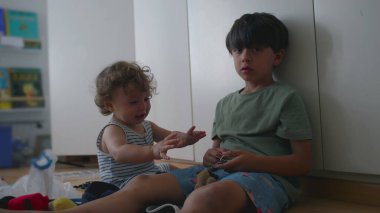 Büyük kardeş, bebek kardeşlerin kafasının yanında anahtarlık oyuncağını sallarken yeni yürümeye başlayan ördekler ve kalkanlar arka planda kitaplarla birlikte çocuk odasında yerde oturuyorlar.