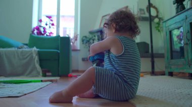 Küçük çocuk içeride oturmuş, mavi oyuncak robotu çeviriyor. Odaklanıyor ve merak ediyor. Etrafı rahat mobilyalarla çevrili ve pencereden içeri ışık giriyor.