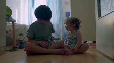 İki küçük kardeş, tahta zeminde güneşli yatak odasında birlikte oynuyorlar. Bebek, oyuncaklarla ve gün ışığıyla çevrili büyük çocuğu sıcak bir şekilde kucaklıyor.