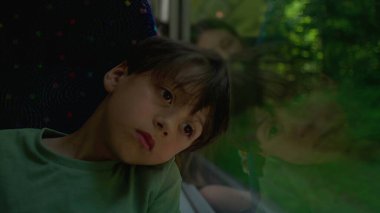 Genç çocuk uzak bir ifadeyle başını cama yaslar, yüzle harmanlanmış orman yansıması, yolculuk sırasında düşünceli bir ruh hali, sessiz bir yolculuk anı