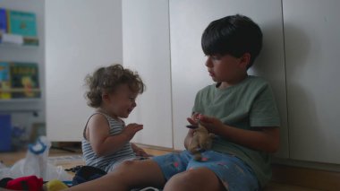 Büyük kardeş, bebek kardeşlerin kafasının yanında anahtarlık oyuncağını sallarken yeni yürümeye başlayan ördekler ve kalkanlar arka planda kitaplarla birlikte çocuk odasında yerde oturuyorlar.