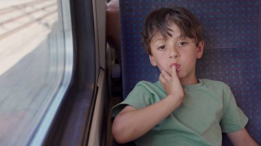 Trenin penceresinde oturan çocuk, el ele tutuşmuş, yolculuk sırasında düşünceli bir ifadeyle dışarı bakıyor, samimi çocukluk anı yansıması ve durgunluk.