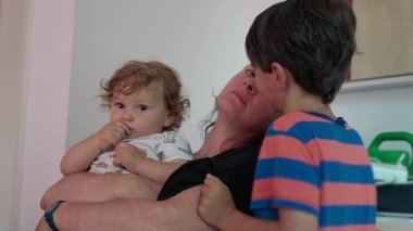 Anne kucağında bebek ve yanaktan nazikçe öpüşürken büyük çocuk yanında duruyor. Evin içinde özgün anne şefkati ve aile şefkati sahnesi.