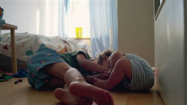 Yürüyen çocuk, büyük kardeşinin yatıp, yatak odasında oynamasını seyrediyor. Camdan gün ışığı akıyor. Etrafa saçılan renkli oyuncaklar.